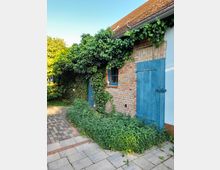 Fassade eines Gebäudes mit roten Ziegeln und blau gestrichenen Holztüren, umgeben von dichter Bewachsung mit Kletterpflanzen und Sträuchern. Davor befindet sich ein gepflasterter Weg und ein kleiner Gartenstreifen.