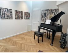 Ein moderner Innenraum mit Fischgrät-Holzboden, einem schwarzen Flügel mit geöffnetem Deckel und Noten auf dem Notenständer. An der weißen Wand hängen vier abstrakte Gemälde, und rechts steht eine skulpturähnliche Figur im Bärenstil.