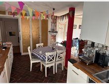 Ein gemütlicher Essbereich mit weiß lackierten Holzmöbeln, einem runden Tisch mit kariertem Tischtuch und einer dekorativen Kristalllampe. Im Hintergrund befinden sich bunte Wimpelgirlanden, eine goldene Schrankfront und eine helle offene Küche mit Arbeitsplatte aus Holz sowie einer Espressomaschine.