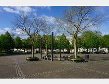 Ein öffentlicher Platz mit gepflasterter Oberfläche, mehreren Bäumen, schlichten Sitzbänken, modernen Metallpfosten und im Hintergrund parkenden Autos sowie Wohnhäusern. Die Umgebung ist von Grünflächen und Laubbäumen umgeben.