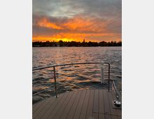 Blick von einem Bootsdeck auf einen See bei Sonnenuntergang, mit leicht welligem Wasser und einer leuchtend orangefarbenen Wolkendecke am Himmel. Am Ufer sind Bäume und vereinzelte Gebäude zu erkennen.