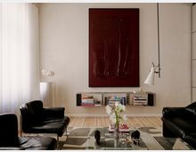 Modernes Wohnzimmer mit hellen Wänden, großformatigem dunkelrotem Wandbild und schwebendem Regal mit Büchern. Schwarze Ledersessel und ein Sofa stehen um einen niedrigen Tisch mit Vase und Deko, neben einer weißen Hängelampe und bodentiefen Vorhängen.