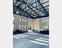 Innenraum einer industriellen Halle mit sichtbaren Stahlträgern an der Decke, großen rechteckigen Fenstern und einer Türöffnung, die nach draußen führt. Im Raum stehen ein schwarzes Auto und ein kleines, flaches Fahrzeug, vermutlich ein Anhänger.