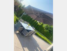 Moderne Garten- oder Terrassenfläche mit großen Steinplatten, einem rechteckigen Glastisch und mehreren geflochtenen Stühlen. Daneben steht ein großer, seitlich montierter Sonnenschirm; im Hintergrund sind hohe Hecken und Bäume zu sehen.