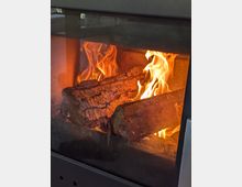 Nahaufnahme eines Holzofens mit brennenden Holzscheiten hinter einer Glasscheibe. Die Flammen lodern hell, und die Oberfläche der Holzscheite ist deutlich sichtbar.