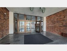 Eingangsbereich eines Gebäudes mit Backsteinwänden, grauem Steinboden und Rasterdecke mit Deckenleuchten. Im Zentrum stehen große verglaste Türen und Fensterrahmen, die in eine helle Lobby mit weiteren Glasfronten führen.