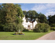 Historisches Gebäude in hellem Stil mit verzierten Fassaden, umgeben von einer gepflegten Parklandschaft mit Blumenbeeten, Bäumen und einem geschwungenen Kiesweg im Vordergrund.