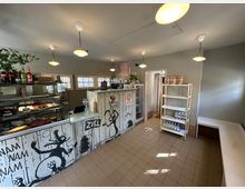 Ein kleiner, heller Innenraum einer Bäckerei oder eines Cafés mit einer Verkaufstheke und Glasvitrine, gefüllt mit verschiedenen Backwaren. Die Thekenfront ist mit Comic-Motiven bemalt, rechts stehen Regale mit Produkten, und an der Decke hängen mehrere runde Deckenlampen.