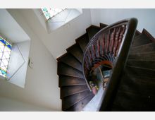Holztreppe in einer Wendelform mit dunkelbraunen Stufen und Balustrade, umgeben von weißen Wänden. Zwei Fenster mit buntem Bleiglas lassen Licht in den engen Treppenbereich.