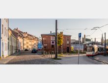 Straßenszene in einer urbanen Umgebung mit Kopfsteinpflasterstraße und farbigen Wohnhäusern auf der linken Seite. In der Mitte ein rotes Backsteingebäude, rechts ein Straßenbahnwagen auf Schienen vor einem Hintergrund mit industriellen Gebäuden und Schornsteinen.