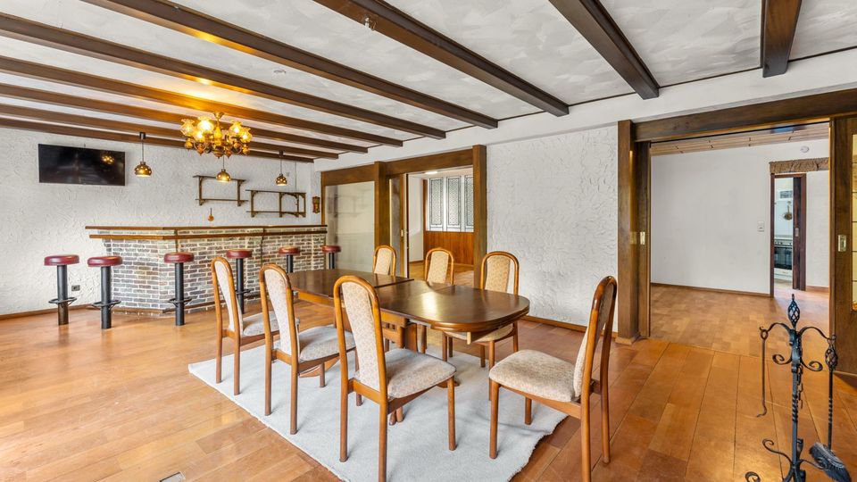 Großer Ess- und Barbereich mit Holzbalkendecke, Holzboden und weiß verputzten Wänden. In der Mitte steht ein Esstisch mit Stühlen auf einem Teppich; im Hintergrund eine gemauerte Theke mit Barhockern, darüber Hängeleuchten und Regale, daneben breite Durchgänge in angrenzende Räume.