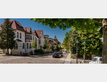 Straßenszene mit historischen Stadthäusern, teils mit Fachwerkelementen und roten Ziegeldächern, entlang einer gepflasterten Straße. Im Vordergrund stehen Bäume, ein Straßenschild mit der Aufschrift "Erich-Mühsam-Straße" sowie parkende Autos.