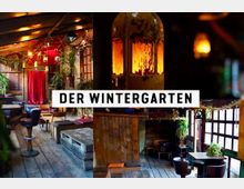 Gemütlicher Innenraum eines Lokals namens „Der Wintergarten“ mit Holzböden, verschiedenen Sitzgelegenheiten aus Holz und Leder und einer warmen Beleuchtung. Pflanzen, Holzdekorationen und rote Vorhänge verleihen dem Raum eine rustikale und einladende Atmosphäre.