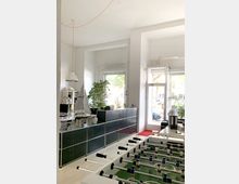 Ein modernes Büro mit einer Schreibtischfläche und einer dunklen Schrankwand vor großen Fenstern, die Tageslicht hereinlassen. Im Vordergrund steht ein Tischkicker, und im Hintergrund sind grüne Pflanzen sowie ein Segelbootmodell zu sehen.