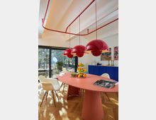 Esszimmer mit modernem Design, featuring einem großen ovalen Tisch in rosa Farbe, darüber drei rote, runde Hängelampen. Im Hintergrund ein blaues Sideboard mit Dekorationen, große Glasfenster mit Blick auf einen Garten, und der Boden ist aus hellem Holzparkett.