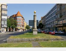 Straßenkreuzung in einer Stadt mit einer historischen Steinsäule mit goldener Kugel auf einem begrünten Mittelstreifen. Umgeben sind die Straßen von mehrstöckigen Wohn- und Geschäftshäusern mit verschiedenen Fassadenstilen, Autos und Geschäften.
