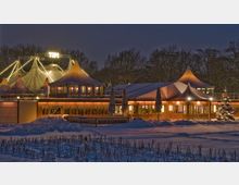 Winterliche Abendaufnahme eines beleuchteten Veranstaltungszelts mit spitzen Dachformen und umliegendem Restaurantbereich. Im Vordergrund liegt eine schneebedeckte Fläche, im Hintergrund sind Bäume zu erkennen.
