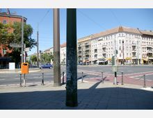 Kreuzung im urbanen Raum mit mehrgeschossigen Wohnhäusern im Hintergrund, Straßenlaternen und Verkehrszeichen, darunter ein Straßenschild mit der Aufschrift „Prenzlauer Allee“. Im Vordergrund ein Gehweg, ein Mülleimer und mehrere Kettenpfosten.