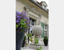 Ein historisches Gebäude mit heller Fassade und grünen Fensterläden, im Vordergrund ein Blumentopf mit lila Blumen; daneben steht eine steinerne Kugel auf einem Sockel als Dekoration.