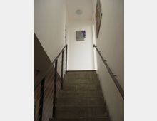 Betontreppe in einem modernen Innenraum, flankiert von weißen Wänden und einem dunklen Geländer mit Metallstreben. An der Wand am oberen Ende der Treppe hängt ein farbiges Kunstwerk, darüber ist eine kleine Deckenleuchte.