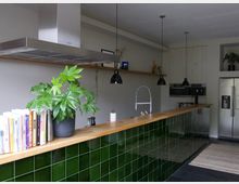 Moderne Küche mit einer langen Arbeitsfläche aus Holz, deren Vorderseite mit glänzenden, dunkelgrünen Fliesen verkleidet ist. Links befinden sich Kochbücher und eine Pflanze, darüber hängen schwarze Pendelleuchten, und im Hintergrund sind eingebaute Küchengeräte sowie ein Edelstahl-Kühlschrank zu sehen.