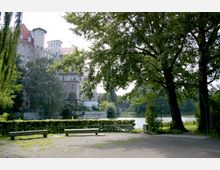 Ein parkartiger Platz mit zwei Bänken im Vordergrund, umgeben von hohen Bäumen und einer metallenen Pergola. Im Hintergrund ist ein großes historisches Gebäude mit Türmen und roten Dächern sowie ein Fluss mit dichter Vegetation zu sehen.