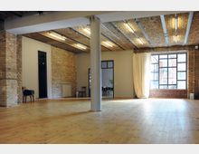 Großer, offener Raum mit heller Holzfußbodenfläche, Backsteinwänden und einer Decke aus freiliegendem Mauerwerk mit integrierter Beleuchtung. Ein großes Fenster mit schwarzen Metallrahmen sowie beigefarbene Vorhänge lassen Tageslicht in den Raum.