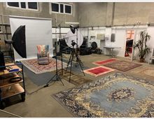 Ein Fotostudio in einem industriell anmutenden Raum mit Betonwänden und hohen Fenstern. Im Vordergrund liegen bunte Teppiche, während im Hintergrund ein Set mit einer weißen Fotoleinwand, einer Lampe, einer Kamera auf einem Stativ und einem dekorativen Sessel steht.