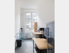 Schmale, helle Büroküche mit großen Sprossenfenstern, grauem Boden und weißen Wänden. Entlang der rechten Seite stehen Arbeitsplatten aus Holz mit Spüle und Kaffeemaschine, links ein Metallregal und ein Abfalleimer.