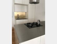 Moderne Küche mit weißen Einbauschränken, einem eingelassenen Edelstahlspülbecken und einem hellen Spritzschutz. Im Vordergrund befindet sich eine Kücheninsel mit einer Edelstahloberfläche, einem integrierten Gasherd und einer Dunstabzugshaube darüber.