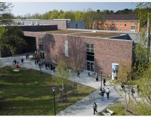 Backsteinbau eines modernen Gebäudes mit Flachdach und großen Fenstern, umgeben von einer Grünfläche und Bäumen. Im Vordergrund befinden sich ein gepflasterter Gehweg, Fahrradständer und mehrere Personen, die das Gelände betreten oder verlassen.