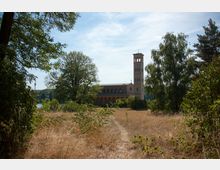 Kirche im neoromanischen Stil mit einem hohen Glockenturm, umgeben von Bäumen und einer trockenen Wiesenlandschaft, im Hintergrund ein See und bewaldete Hügel.