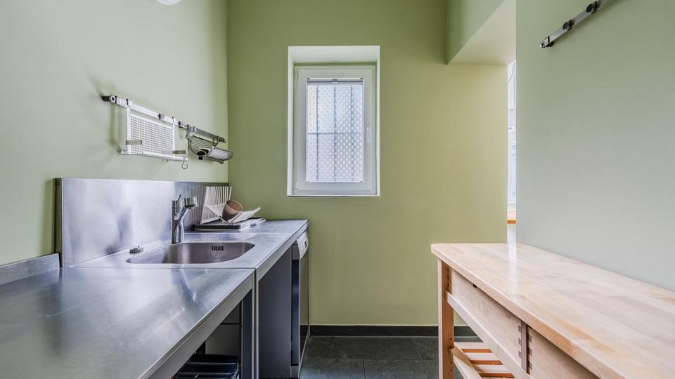 Schmale Küche mit hellgrünen Wänden, Edelstahl-Arbeitsflächen und Spülbecken an der linken Seite sowie einem großen Holz-Arbeitstisch rechts. Am Ende des Raums befindet sich ein kleines Fenster; der Boden ist mit dunklen Fliesen ausgelegt.