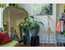 Cozy apartment studio corner with a large floor mirror draped in ivy, potted leafy plants on dark stands, and an abstract painting on a white wall. A wooden easel stands beside a small clothing rack, with a sofa edge and record player visible in the foreground.