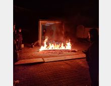 Ein brennendes Feuer auf dem Boden vor einem geöffneten Frachtcontainer bei Nacht, beleuchtet von den Flammen. Personen stehen in der Nähe und beobachten die Szene, während im Inneren des Containers zwei weitere Personen sichtbar sind.