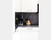 Moderne Küchenzeile mit weißen Schränken und grauer Arbeitsplatte. Auf der Arbeitsfläche befinden sich eine schwarze Kaffeemaschine und ein Messerblock aus Holz mit mehreren Messern.