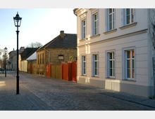 Pflastersteinstraße in einer ruhigen Wohngegend mit historischen Gebäuden, darunter ein modernes cremefarbenes Gebäude mit weißen Fenstern und ein altes Backsteinhaus mit rotem Tor. Entlang der Straße stehen schwarze Laternen, und im Hintergrund sind weitere Häuser sichtbar.