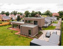 Moderne, kubische Wohnhausarchitektur mit Flachdächern und brauner Fassade, umgeben von einem gepflegten Garten. Im Vordergrund eine gepflasterte Einfahrt mit geparkten Autos, im Hintergrund eine Nachbarschaft mit Einfamilienhäusern und Bäumen.