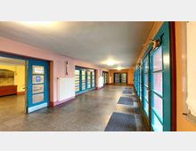 Ein Flur mit hell gestrichenen Wänden, mehreren blauen Türen mit Glaseinsätzen auf beiden Seiten und grauem Steinboden. Beleuchtung erfolgt durch Deckenlampen, an den Wänden befinden sich Heizkörper und Wandlampen.