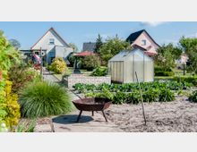 Ein Garten mit Gemüsebeeten, einem kleinen Gewächshaus und einem gemauerten Hochbeet. Im Hintergrund sind Einfamilienhäuser, Büsche, Obstbäume und ein Wäscheplatz zu sehen.