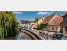 Straßenszene in einem kleinen Ort entlang eines Kanals mit beidseitigem Geländer; links eine große Weide, rechts eine Reihe von Häusern mit farbigen Fassaden und roten Dächern. Geparkte Autos stehen entlang der Straße, blauer Himmel mit vereinzelten weißen Wolken im Hintergrund.