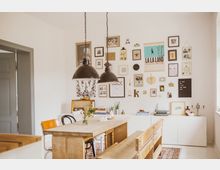 Esszimmer mit hellen Holzmöbeln, darunter ein großer Esstisch, gemischte Stühle und eine Holzbank. An der weißen Wand hängen zahlreiche Bilder und Dekorationen in unterschiedlichen Rahmen; darüber befinden sich zwei schwarze Hängelampen. Ein weißer Sideboard steht unter den Wanddekorationen, dekoriert mit Büchern, einer Schreibmaschine und kleinen Pflanzen.