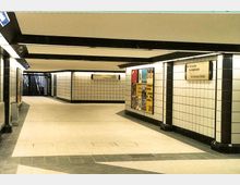 Ein unterirdischer U-Bahn-Gang mit hellen, weiß gefliesten Wänden, schwarzen Rahmenakzenten und einem grauen Fliesenboden. An den Wänden befinden sich Wegweiser und Werbeplakate, im Hintergrund sind Treppen zu sehen.