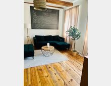 Modernes Wohnzimmer mit dunkelgrünem Samt-Ecksofa auf hellem Teppich, rundem Couchtisch mit Holzplatte und rustikalem Dielenboden mit sichtbaren Holzbalken an der Decke. An der Wand hängt eine große Periodensystem-Tafel, neben bodenlangen Vorhängen steht eine Topfpflanze.