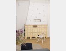 Ein heller Innenraum mit einem cremefarbenen Kachelofen an einer weißen Wand, verziert mit runden Öffnungen und einer kleinen rechteckigen Klappe. Im Vordergrund steht ein weißer Klappstuhl, während der Boden teils aus beigem Holz und teils aus grauem Material besteht.