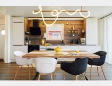 Moderne Küche mit Holzakzenten, ausgestattet mit weißen Schränken, einer dunklen Arbeitsplatte und einem zentralen Esstisch aus Holz mit Stühlen in Weiß und Schwarz. Im Hintergrund befindet sich eine Wand mit Holzverkleidung, ein Fenster, offene Regale mit goldenen Accessoires und eine auffällige, geschwungene Hängelampe.