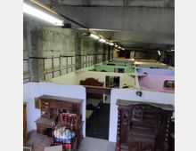 Innenansicht eines Lagerhauses mit hohen Betonwänden und einer offenen Betondecke. Der Raum ist in kleine Abteile mit bunten Wänden unterteilt, die antike Möbelstücke wie Schränke und Tische enthalten.