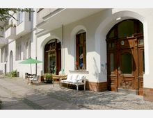 Straßenszene mit einer Altbaufassade, definiert durch mehrere hohe weiße Bögen und braune Holztüren. Vor einem Schaufenster steht eine Sitzbank mit weißen Kissen und ein Tisch mit grünem Sonnenschirm auf einem gepflasterten Gehweg.