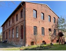 Backstein-Gebäude mit zwei Etagen, hohen rechteckigen und bogenförmigen Fenstern, darunter einige mit kleinen Sprossen. Das Gebäude steht in einer grünen, offenen Umgebung mit Rasen und Büschen, und im Vordergrund ist eine moderne Metallskulptur sichtbar.