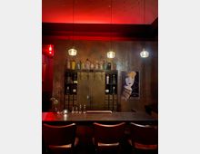 Eine stilvolle Bar mit dunkler Theke und Holzhockern, umgeben von warmem roten Licht. Im Hintergrund befinden sich ein Regal mit Glasflaschen, ein großer Metallschrank und ein Retro-Poster an der Wand.
